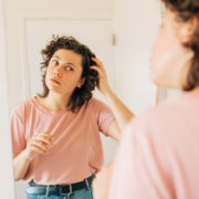 Young woman with curly hair looking at herself in the mirror and combing hair with hand, photographed from behind