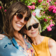 Mature mother and daughter embrace outside in front of bright flowering tree. It is summer, they are both wearing trendy sunglasses and smiling.
