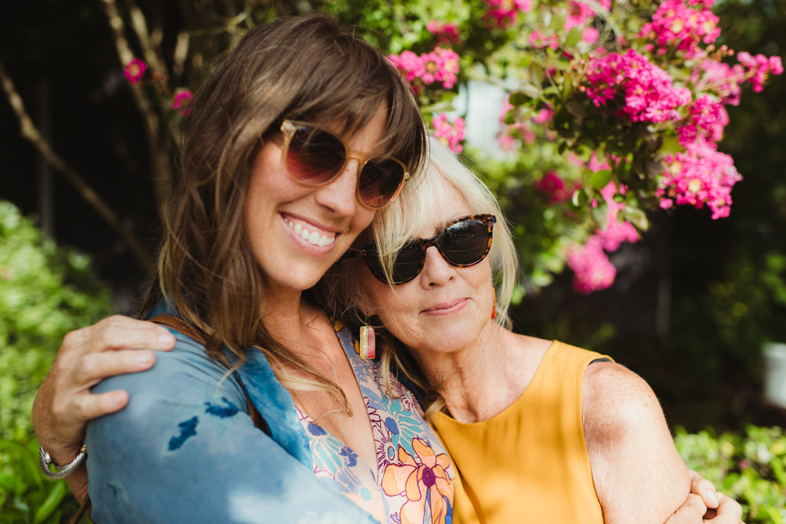 Mature mother and daughter embrace outside in front of bright flowering tree. It is summer, they are both wearing trendy sunglasses and smiling.